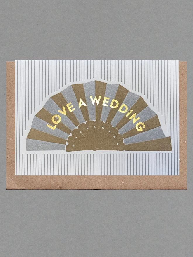 White and silver striped card with silver and gold fan on it and gold text reading 'Love A Wedding' on it with a brown envelope behind