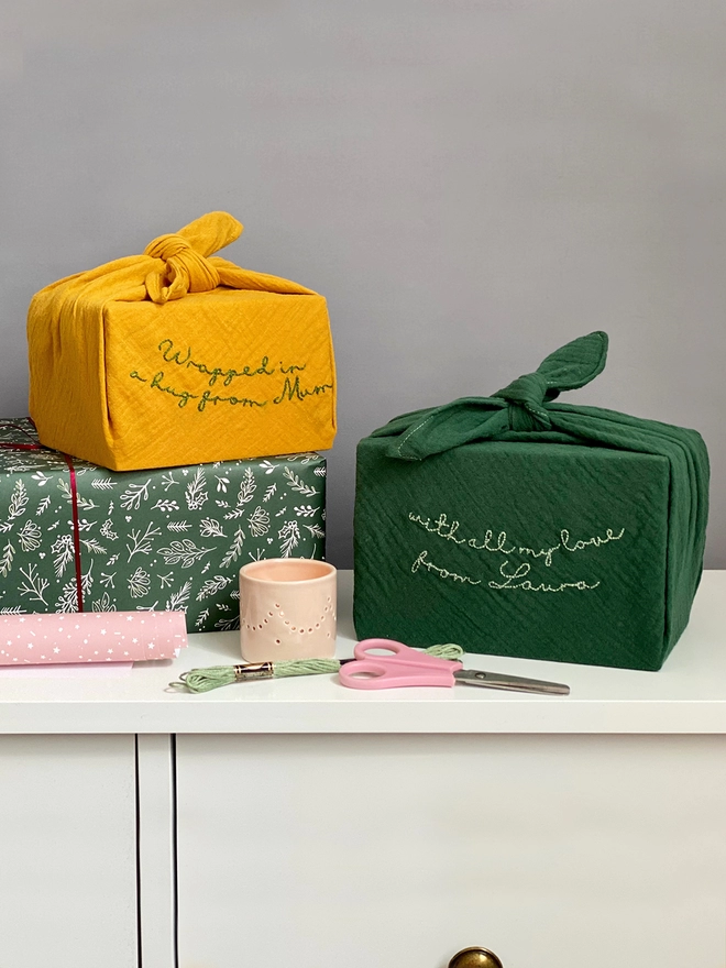 Two gifts wrapped in cotton fabric are stood on a white desk. Each one has been embroidered with a special message.