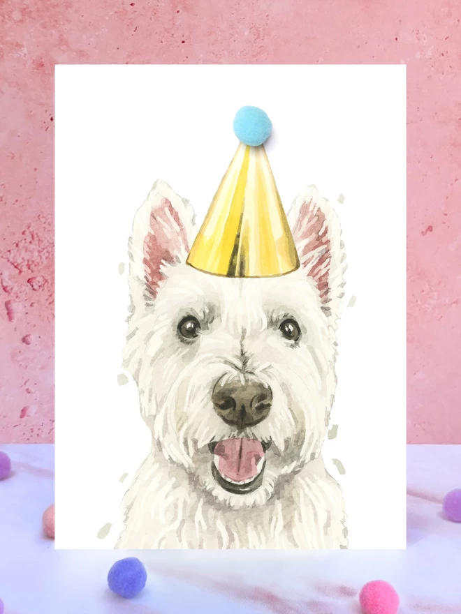 A greeting card featuring a hand painted design of a westie, stood upright on a marble surface surrounded by pompoms. 