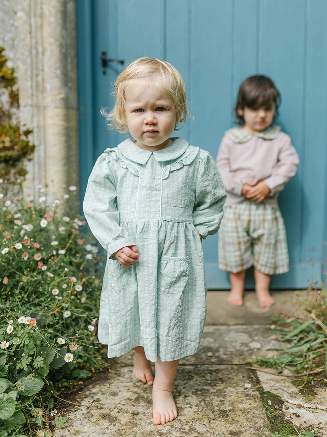 A little girl in a green checked dress with long sleeves and a collar walks in front of a little boy in a brown and green short set