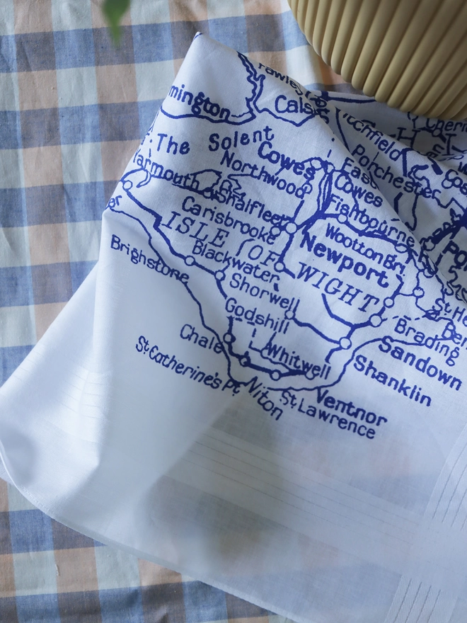A lifestyle photo of a Mr.PS Solent Map hankie printed in blue on a gingham tablecloth with a yellow plant pot