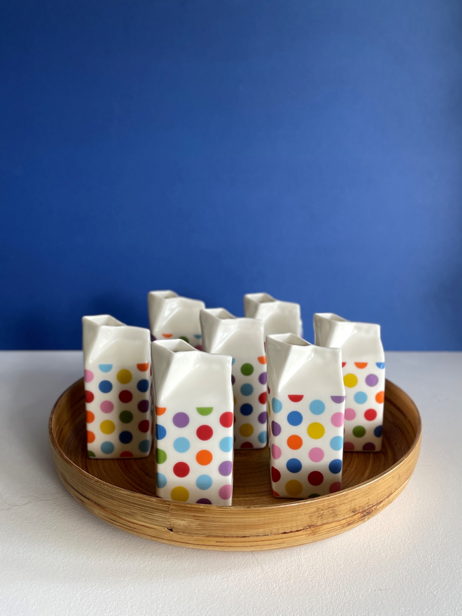 Group of seven colourful polka-dot porcelain milk carton jugs displayed on a round bamboo tray  , on a cobalt blue back ground 