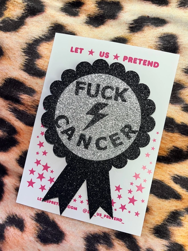 A rosette with a silver disc in the centre featuring the words ‘FUCK CANCER’ and a lightening bolt in black. The scalloped edging and tails are black.