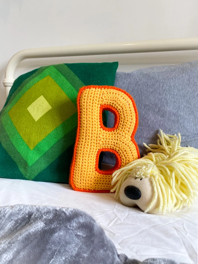 Crocheted cushion shaped like the letter B in Pale Orange and Orange on a childs bed