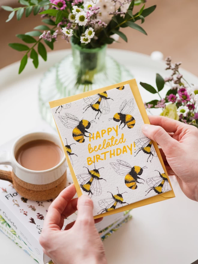 happy belated birthday plantable card held above tabletop with vase of wildflowers