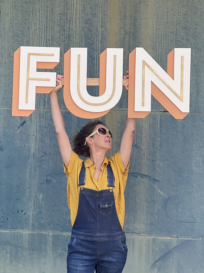 A person wearing dungarees and sunglasses holds a wooden sign reading FUN above their head