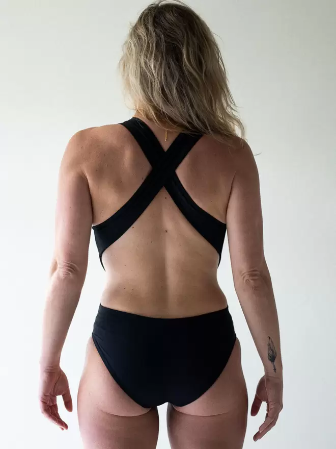 Blonde woman in studio facing away with hair over one shoulder, wearing black Davy J Sustainable Waterwear cutout swimsuit with cross back straps