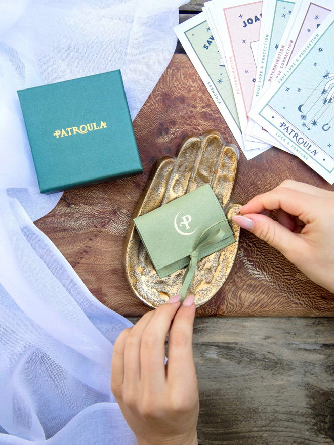 Hands untying a pale green Patroula Jewellery pouch with a giftbox and postcards in the background