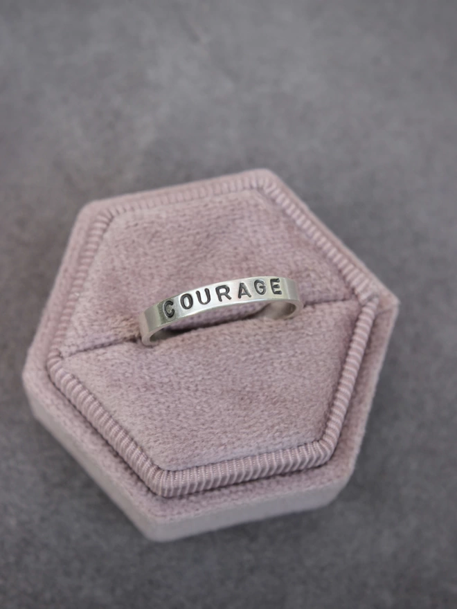 A single silver plain band ring with the word 'courage' stamped in uppercase font, in a hexagonal pink velvet ring box on a lilac background.