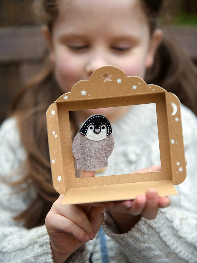 A young girl holds a felt penguin finger puppet on her finger beside a small cardboard puppet theatre.