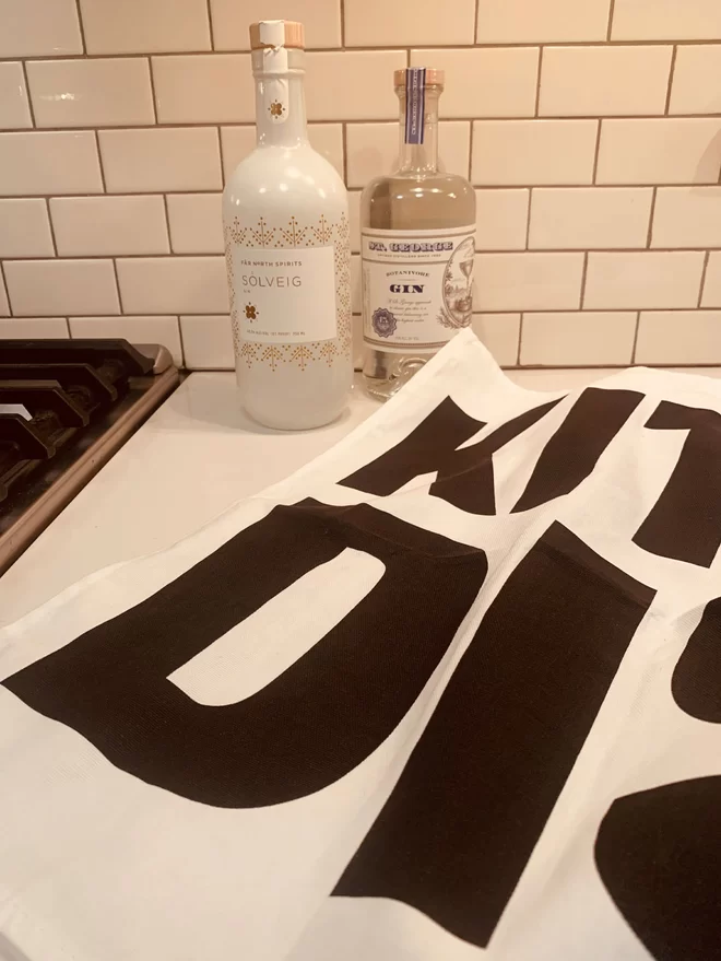 Close up of a few letters of Kitchen Disco black screen printed text on white tea towel sat on kitchen counter in front of a fee bottles of gin