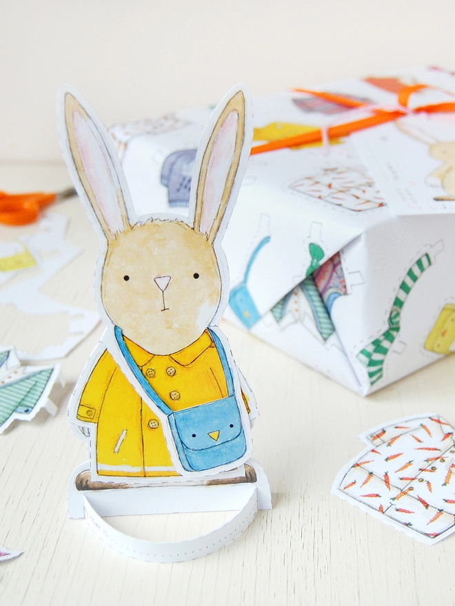 A gift wrapped in white wrapping paper with a hand illustrated outfit design and rabbit paper doll gift tags is beside a cut out rabbit paper doll on a white wooden table.
