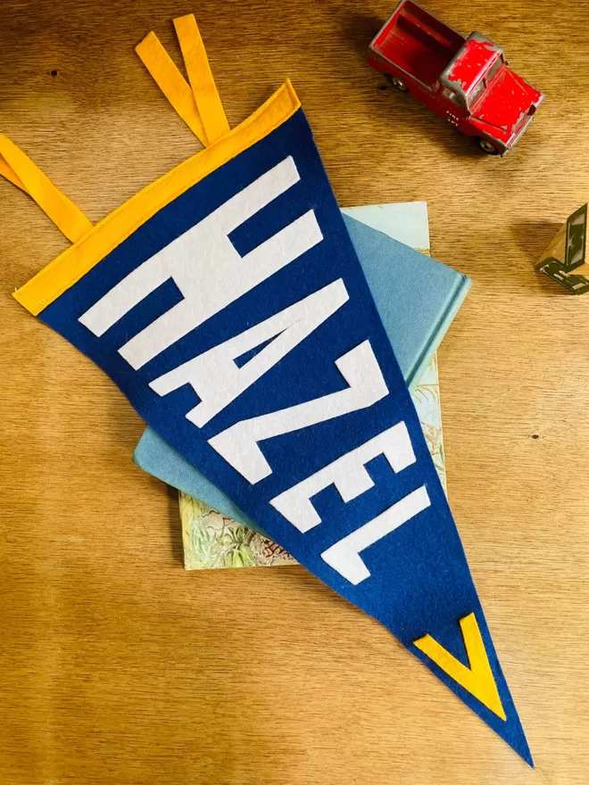 A blue and yellow pennant flag with the name Hazel lying on a eooden table with a toy car.
