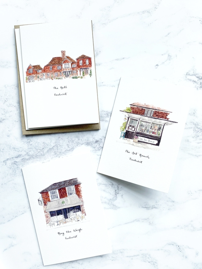 Watercolour illustrated Ticehurst shop front cards on A6 white cards. Featuring five beautiful independent small businesses in Ticehurst, East Sussex - The Bell, Buy the Weigh and The Art Bunch, then Greenfinch and The Old Haberdashery are not pictured. The cards sit slightly open, laid flat with brown kraft paper envelopes underneath. The background is a pale white marble