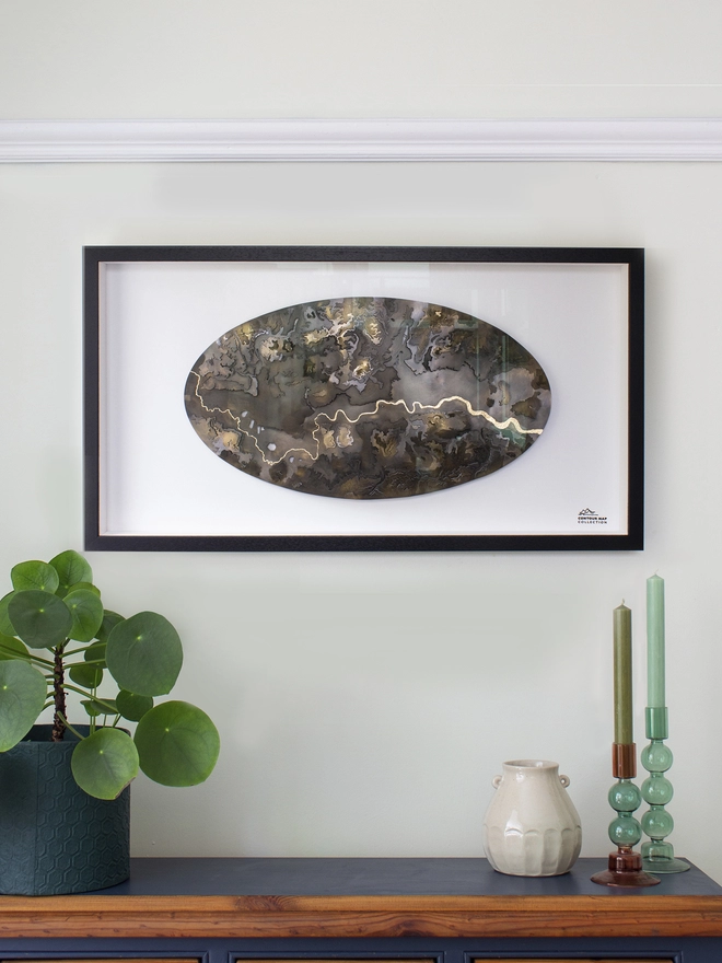 A large oval topographical metal map with a golden river, mounted on a white background with a black frame.