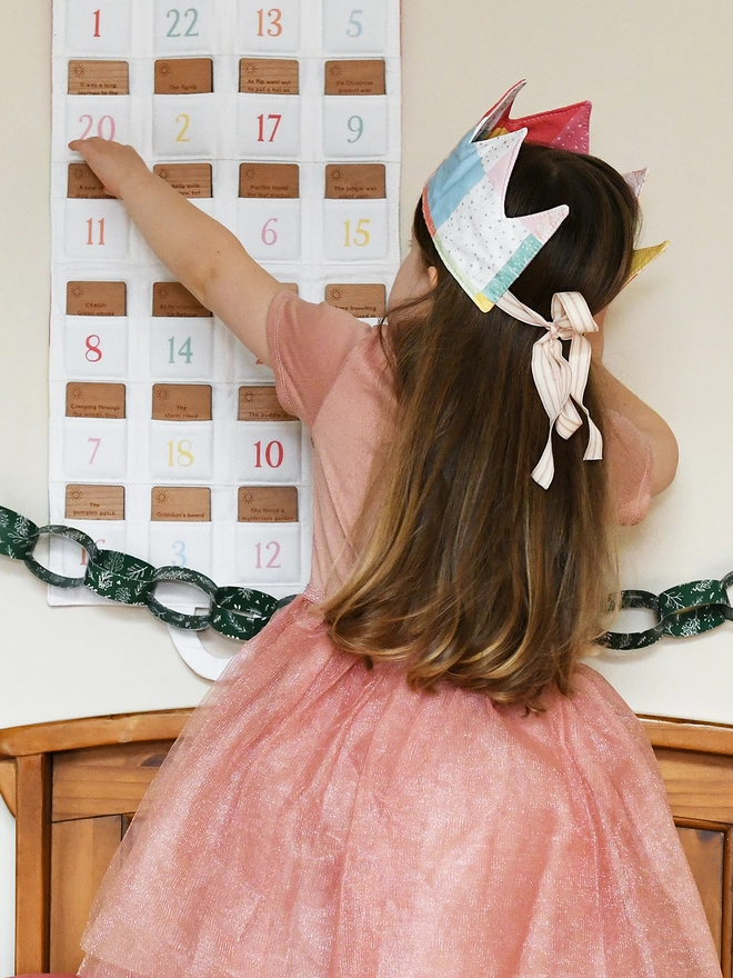 A young girl wearing a pink tutu dress and a pastel patchwork crown has her back to the camera and is looking at a fabric advent calendar.
