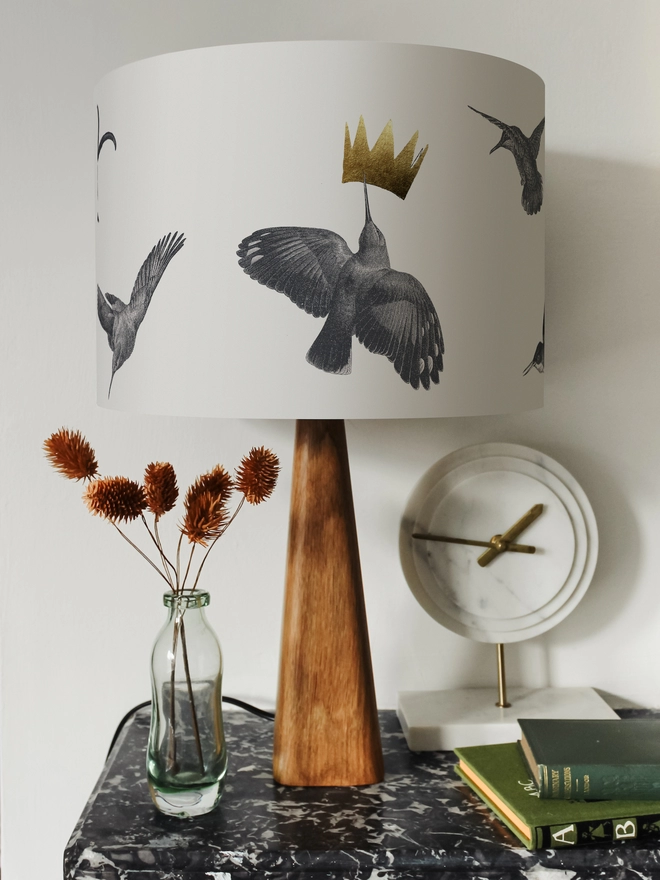 Drum Lampshade featuring hummingbirds on a wooden base on a shelf with books and ornaments