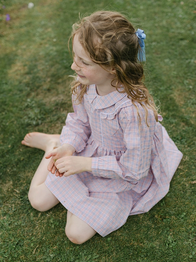 A girl in a pink checked dress sits on a lawn