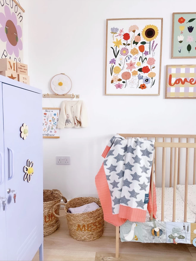 A view of a bright and colourful nursery with a wooden cot. Draped over the side of the cot  is a grey and white star baby blanket with coral pink trim. Modern floral prints line the walls and toys are stacked neatly in wicker baskets on the floor.
