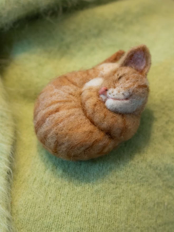 A needle-felted sleeping ginger tabby cat brooch on a green fluffy jumper