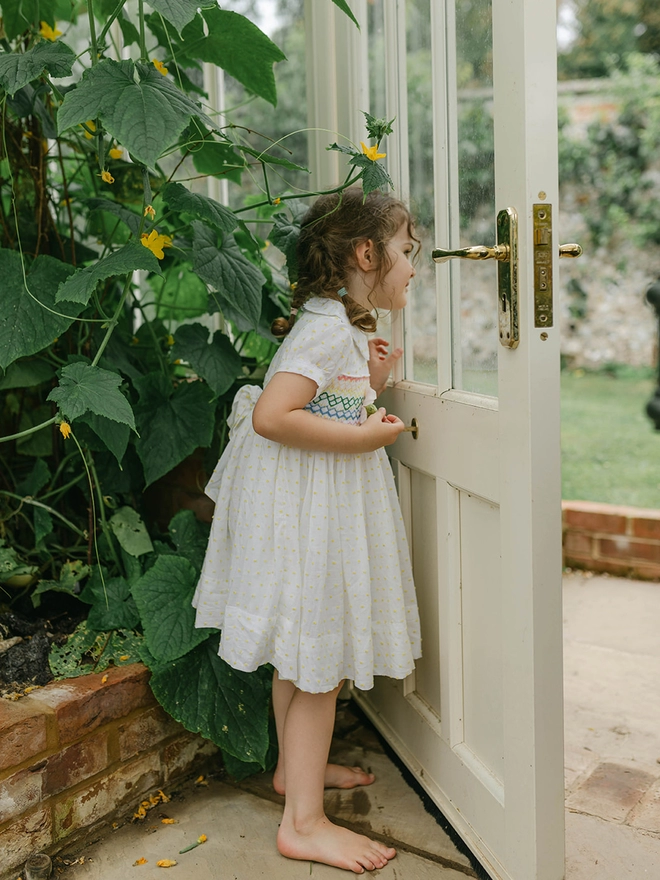 A girl looks out a greenhouse door in a white and yellow swiss dot dress with a peter pan collar and rainbow smocking