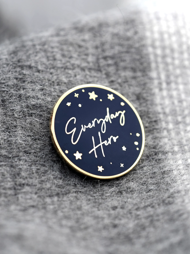 A navy blue and gold enamel pin badge with a gold star design and the words "Everyday Hero" is pinned to a grey blanket.