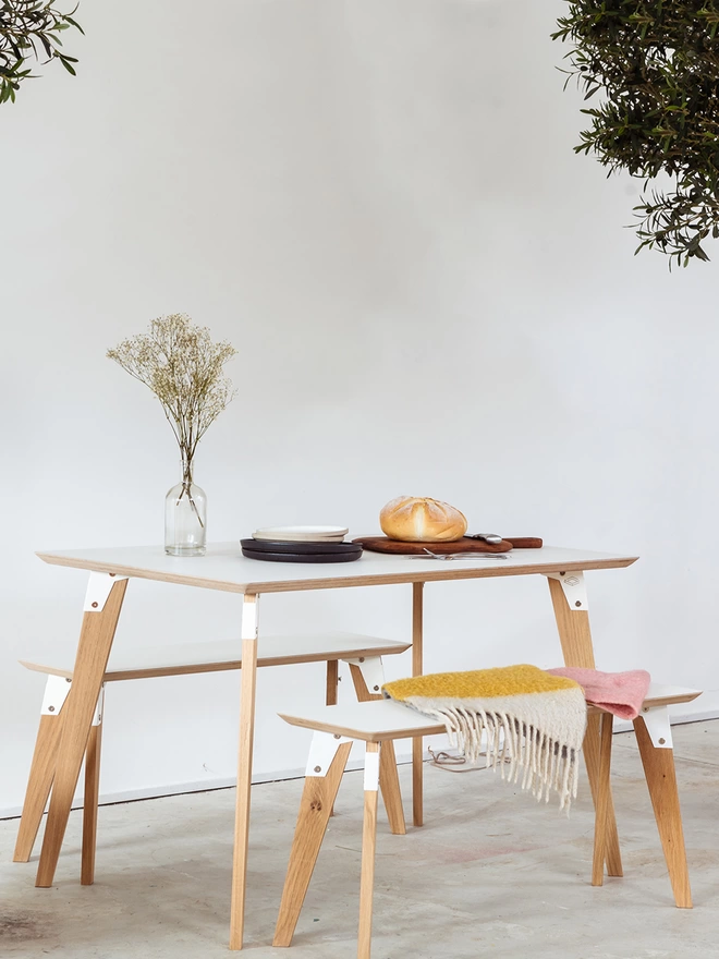 A stylish and minimalist dining set comprising a table and benches with white Fenix top, white coloured steel brackets and solid oak legs, laid for a simple lunch with a loaf of fresh bread