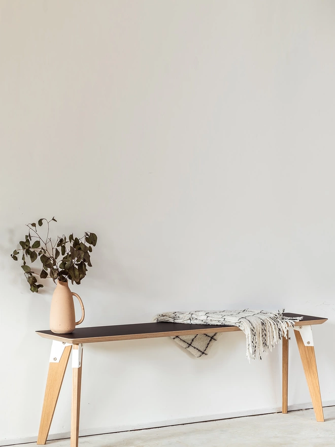 angle view of a stylish and minimalist bench with black Fenix top, white coloured steel brackets and solid oak legs with a throw and vase on it