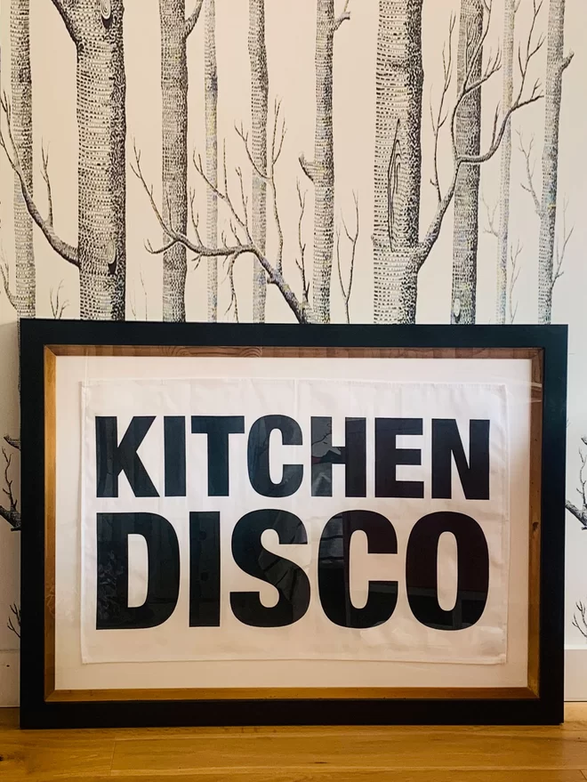 London Drying Kitchen Disco black screen printed text on white tea towel framed in black border frame and sat in front of black lino cut style trees pattern wallpapered wall