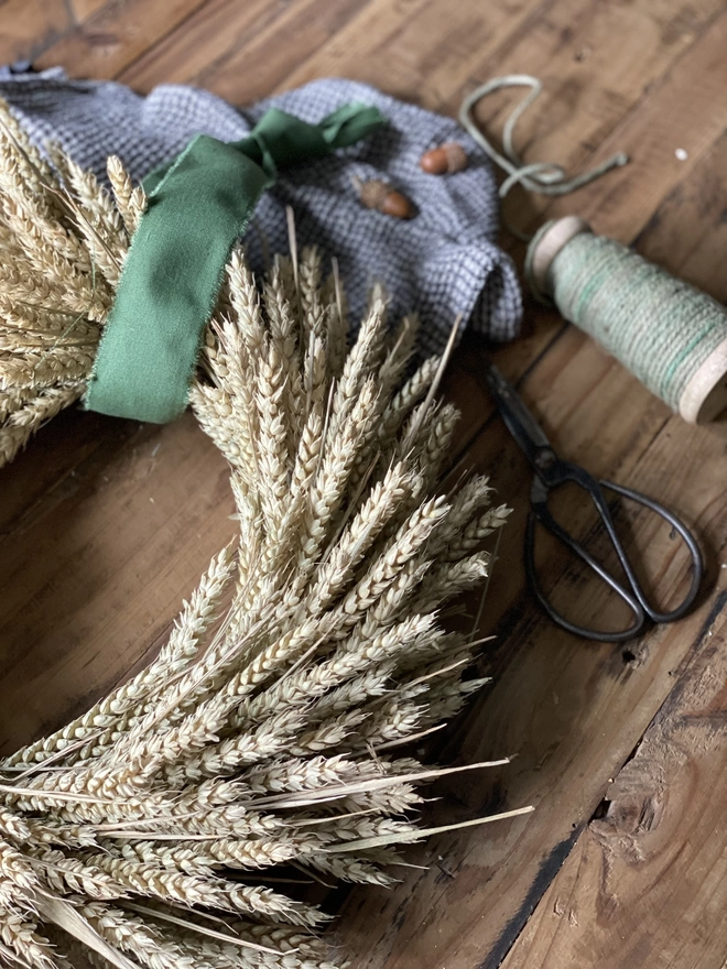 A closer look at the right curve of a handmade September Harvest Wheat Wreath with a sage green ribbon looped around the top, on display with a blue chequered cloth and small autumnal accessories