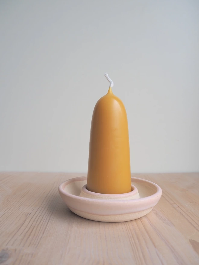 Small stubby beeswax candle in a pale pink candle holder on pale wood surface