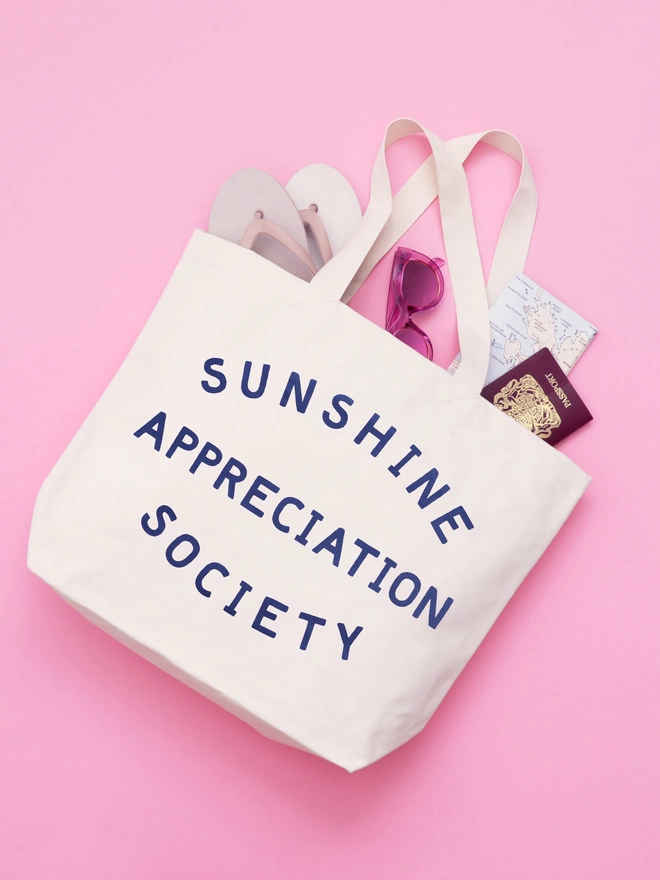 a large natural canvas tote bag bearing the words sunshine appreciation society laying on a pink backdrop with beach items spilling out
