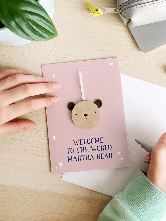 A pink greetings card with a small wooden bear keepsake and the words "Welcome to the world Martha bear" printed on is on a wooden desk. 