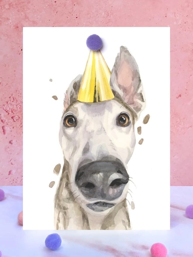 A greeting card featuring a hand painted design of a greyhound, stood upright on a marble surface surrounded by pompoms. 