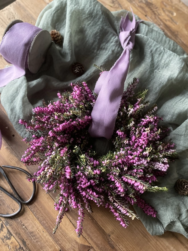 A Petite Wild Heather Wreath in purple and green hues, with a soft lilac ribbon looped through the middle, on display atop a sage green fabric