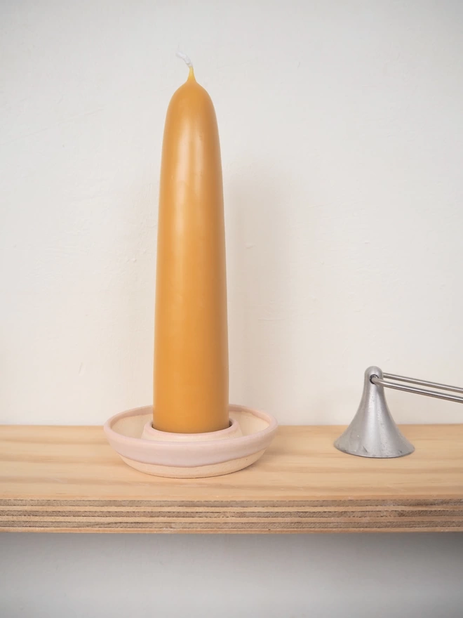 Wooden shelf containing handmade ceramic candle holder with pale pink glaze. In the holder is a giant stubby beeswax candle, next to it a candle snuffer