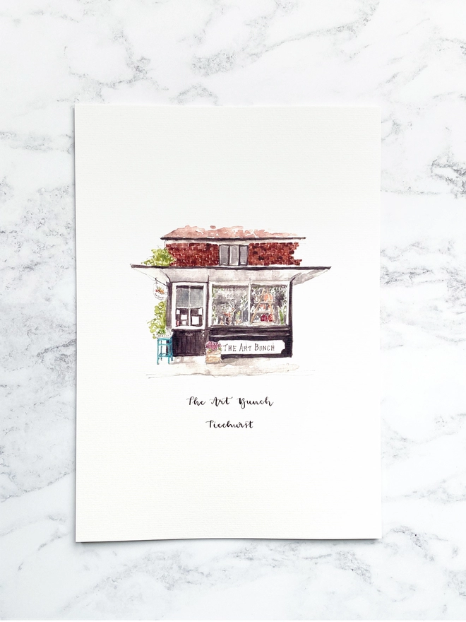 Beautiful watercolour illustration of The Art Bunch florist shop in Ticehurst.  A black and white fronted building with plants outside and in through the large window. The watercolour style is painted with a black pen outline and organic loose style with small details. The print is a small illustration on the centre of a white page and the paper sits on a white marble background.