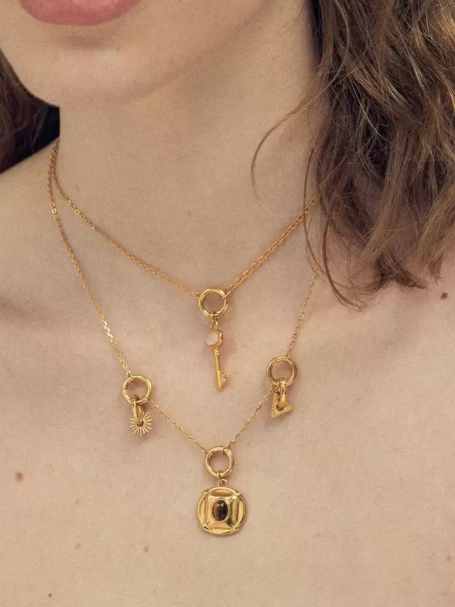 woman wearing two gold necklaces styled with a moonstone key pendant, a tiger's stone medallion and two gold charms