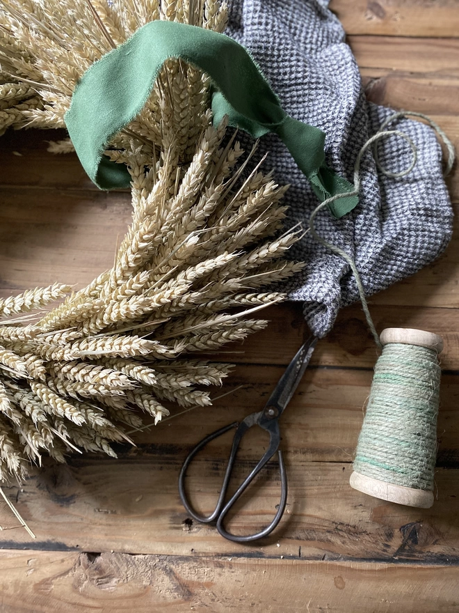 A handmade September Harvest Wheat Wreath with a sage green ribbon looped around the top, on display with a blue chequered cloth, small fabric scissors and a spool of green thread