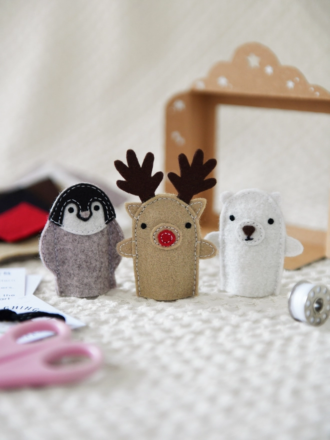 Three felt finger puppets, a reindeer, a penguin, and a polar bear, stand beside a small cardboard puppet theatre on an ivory fabric surface.