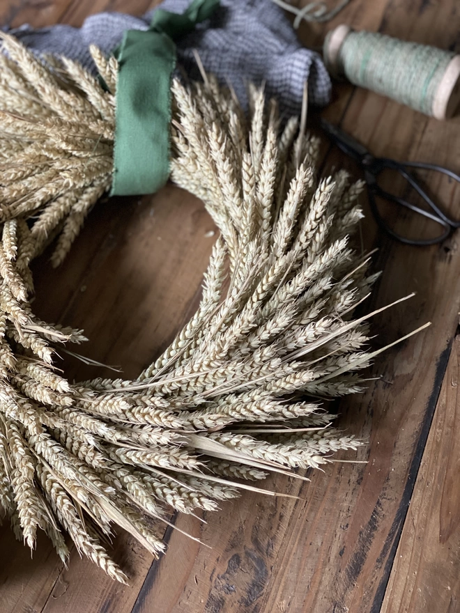 A handmade September Harvest Wheat Wreath with a sage green ribbon looped around the top, on display with a blue chequered cloth and a spool of green thread