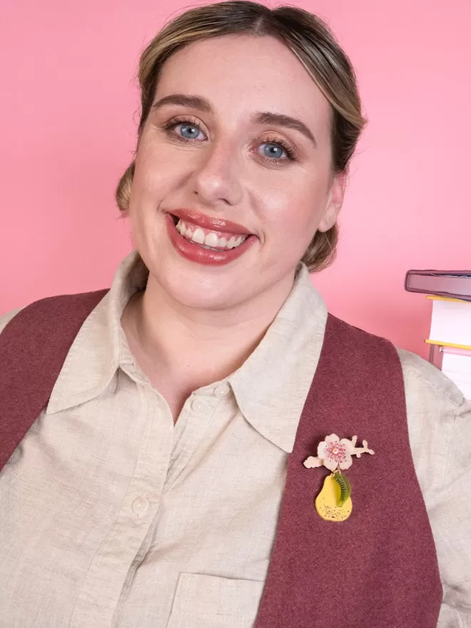 Model is wearing a world book day pear brooch. It is attached to a pink waistcoat worn over a shirt.