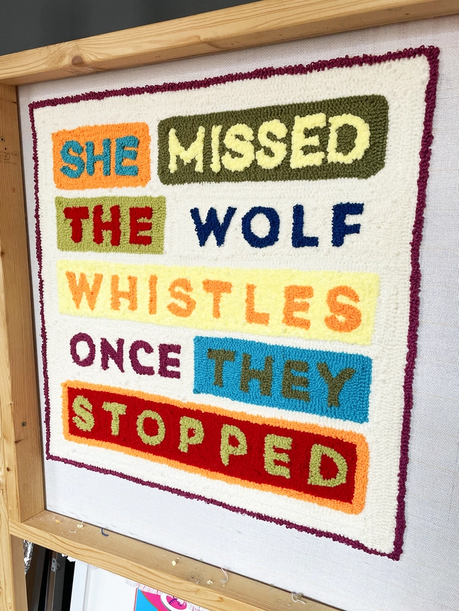 Typographic punch needle textile art "She missed the wolf whistles once they stopped" in bright wool type with coloured blocks in situ on wooden frame