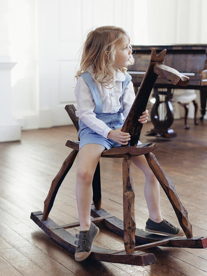 A boy in a white shirt and blue shorts with braces sits on a rocking horse