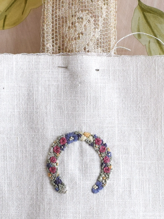 An embroidered Heart, of Lavender Blues and Buttermilk yellow blossoms.  Pinned to a length of vintage lace, with ends pointing down to allow Luck to flow over all who pass by.