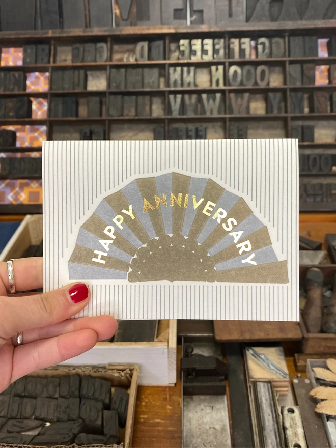 A hand holds up a white card with silver and gold fan with gold text reading 'Happy Anniversary' on silver and white striped card.
