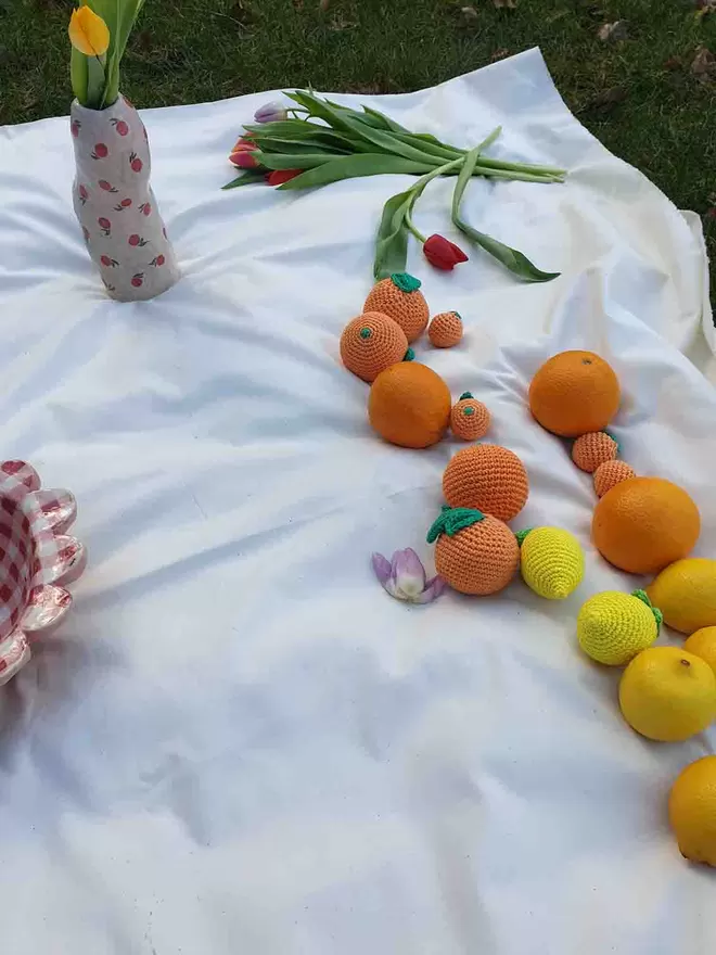 Crochet oranges and lemons and regular oranges and lemons placed on a white cloth with tulips in the background and handmade ceramics.