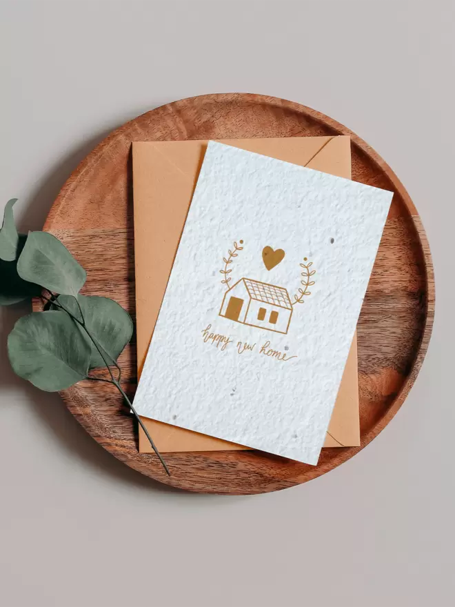 Seed Card with an illustration of a house with a heart and leaves above it with ‘Happy New home’ written beneath on a wooden tray next to a Eucalyptus branch