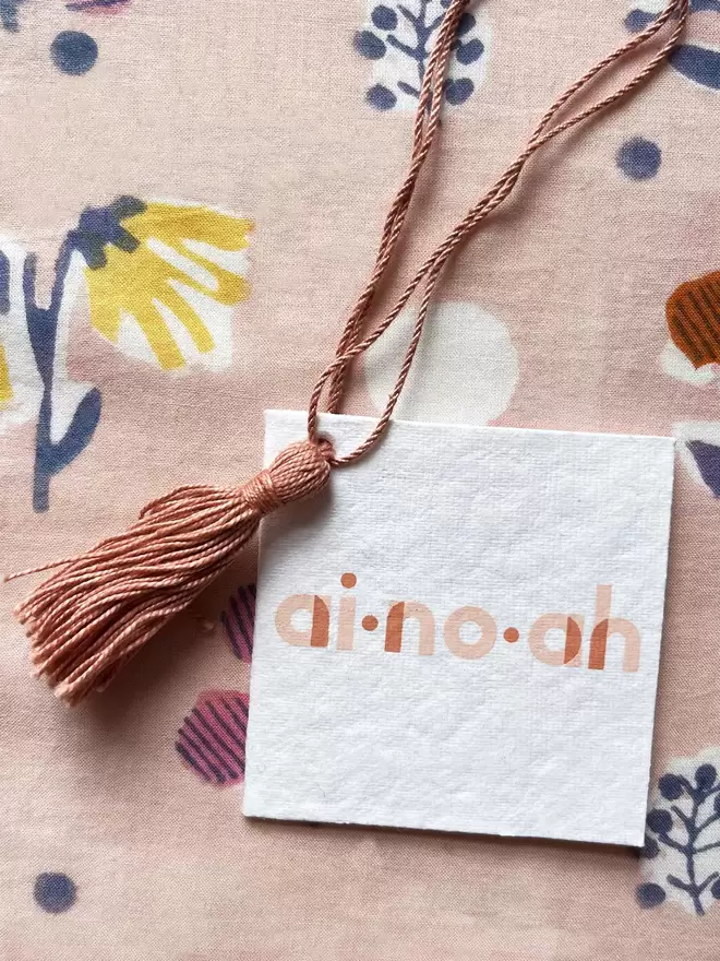 a white hang tag with a printed logo saying ai.no.ah with a pale pink tassel