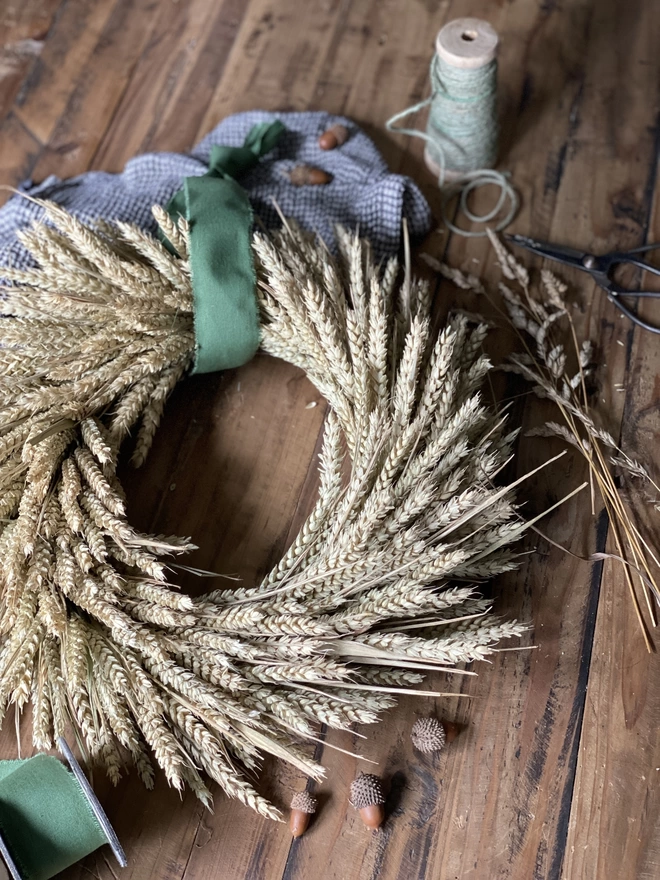 A handmade September Harvest Wheat Wreath with a sage green ribbon looped around the top, on display with a blue chequered cloth, small autumnal accessories and a few strands of loose wheat